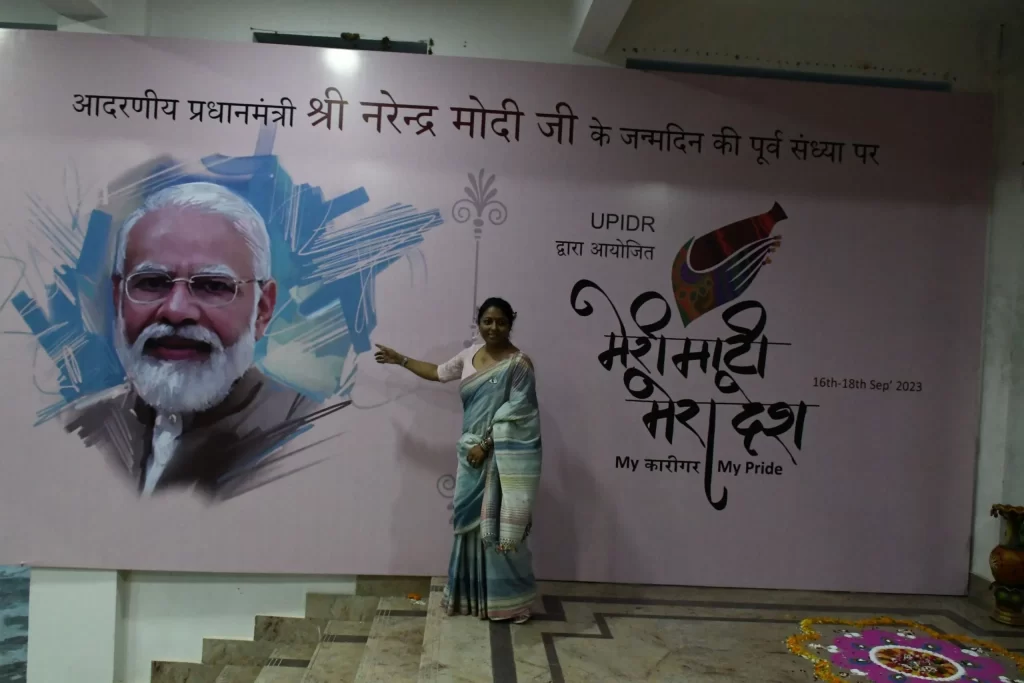 Honorable Kshirpa Shukla standing in front of the poster and pointing to photo of PM Modi ji at the meri mati mera desh event by UPIDR. 